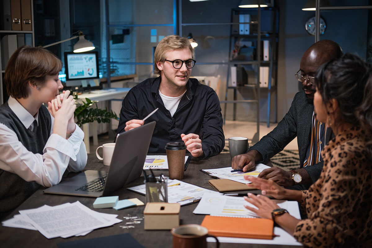 Un groupe de quatre personnes en réunion dans un bureau moderne, discutant autour d'une table avec des ordinateurs portables, des tasses et des documents étalés. L'homme au centre, portant des lunettes, explique quelque chose tandis que les autres l'écoutent attentivement.